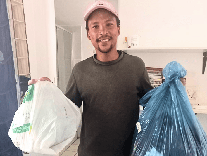 A coloured man stands holding two bags of clothing and smiling broadly. 